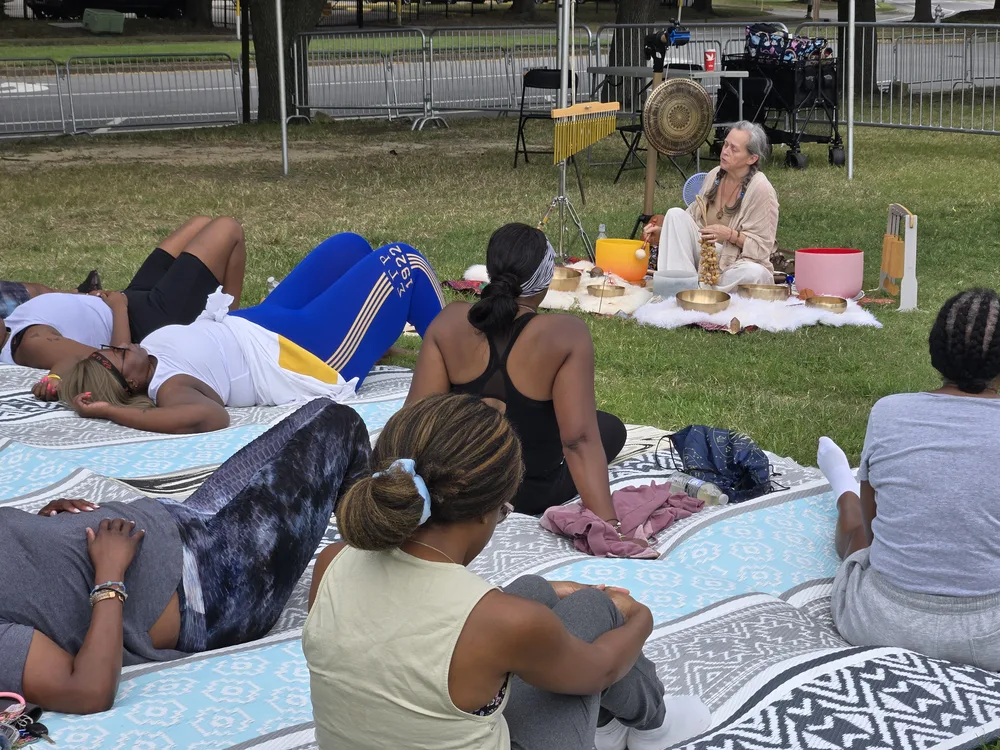 Amy leading an outdoor community sound bath, playing singing bowls and a gong while participants relax on mats on the grass